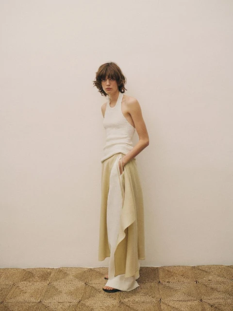A woman wearing a white top and light yellow skirt poses indoors against a neutral wall on a patterned beige carpet.