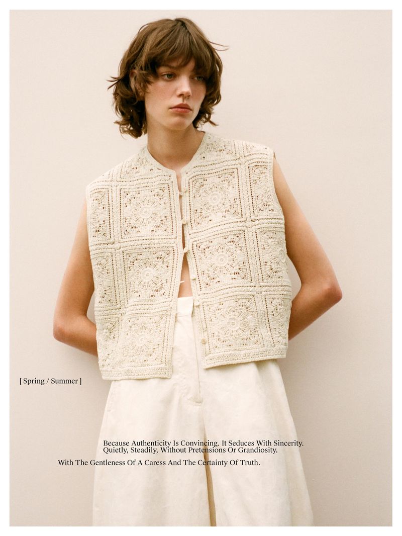 Fashion shot: Young adult woman poses in a cream-colored, square-patterned crochet vest and light-colored pants against a neutral backdrop.