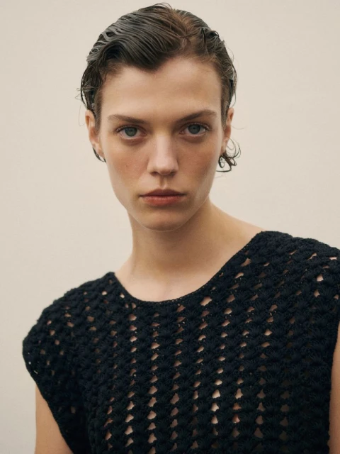 Close-up studio shot of a fair-skinned woman with wet dark hair wearing a dark crocheted top, gazing directly at the camera.