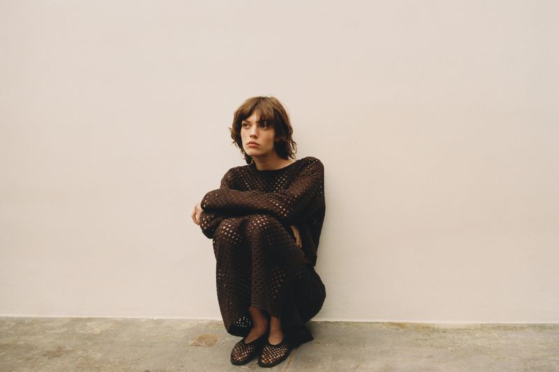 Model wearing a dark brown crochet top and skirt set crouches against a plain neutral studio wall.