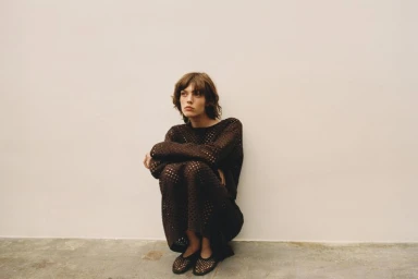 Model wearing a dark brown crochet top and skirt set crouches against a plain neutral studio wall.