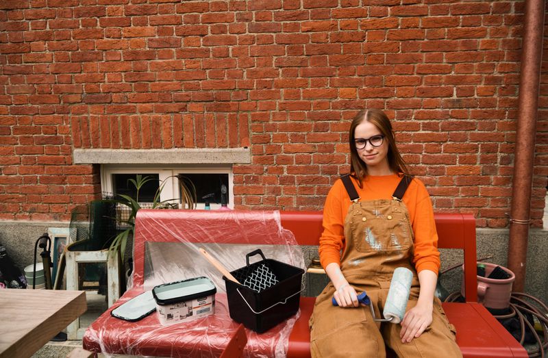 A young woman in overalls sits on a red bench with painting supplies, in front of a brick wall, looking at the camera with a neutral expression.