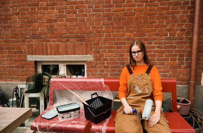 A woman wearing glasses and brown overalls holding a paint roller sitting on a red bench against a brick wall for Fielmann ss24