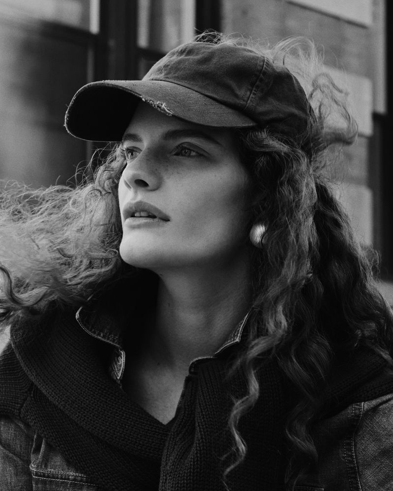 Portrait of an adult female with curly hair wearing a cap, set against a blurred background, shot in black and white with natural lighting.
