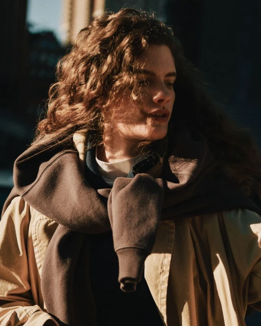 Portrait of a young adult woman with curly brown hair, wearing a tan jacket and brown sweatshirt outside in natural light.