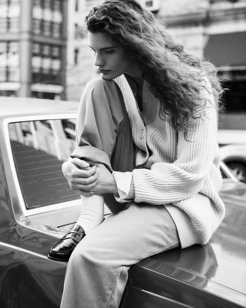 Black and white portrait of a female fashion model sitting on a car hood in casual wear with long curly hair in an urban environment.