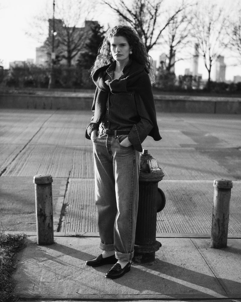 Black and white outdoor portrait of a curly-haired woman in jeans and a shirt, standing on a city sidewalk next to a fire hydrant.