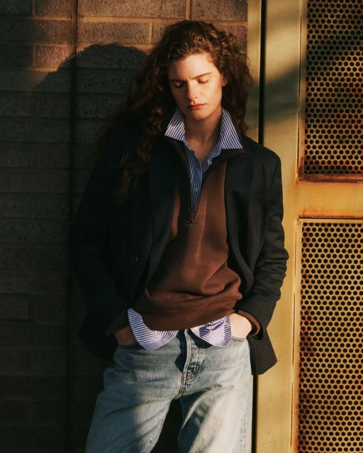 An adult female with curly hair poses in casual fashion, wearing jeans and a blazer, standing in front of a brick wall and metal door.