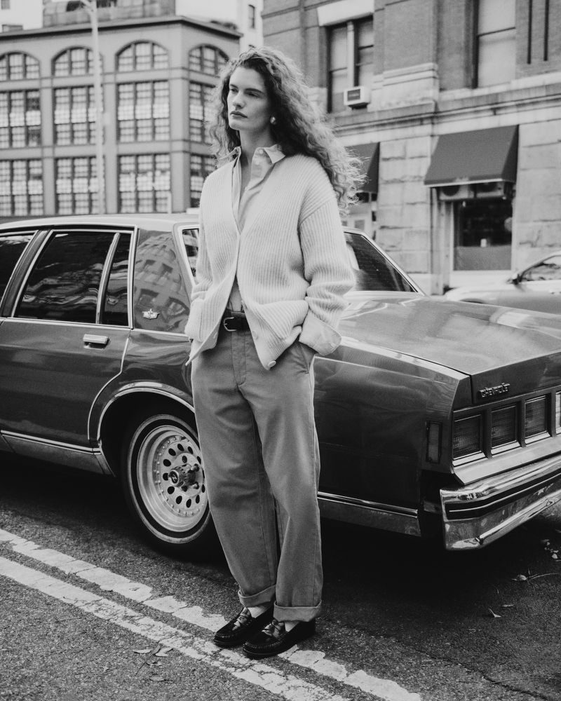 Woman stands in front of a vintage car on a city street, wearing a sweater, button-down shirt, pants, and black shoes in monochrome.
