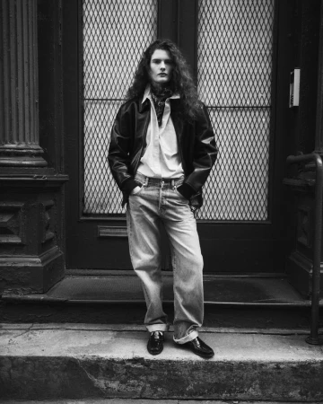 Monochrome portrait of a stylish adult male with long wavy hair in an unbuttoned shirt, leather jacket, jeans and loafers, standing in front of a doorway.