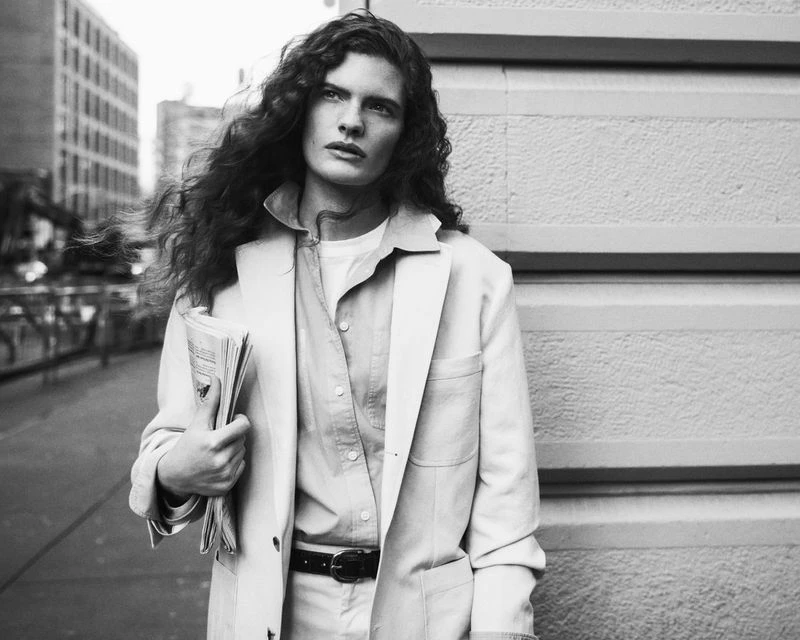 Monochrome portrait of a person with curly hair in a jacket holding a newspaper, standing outside against a neutral-colored building.