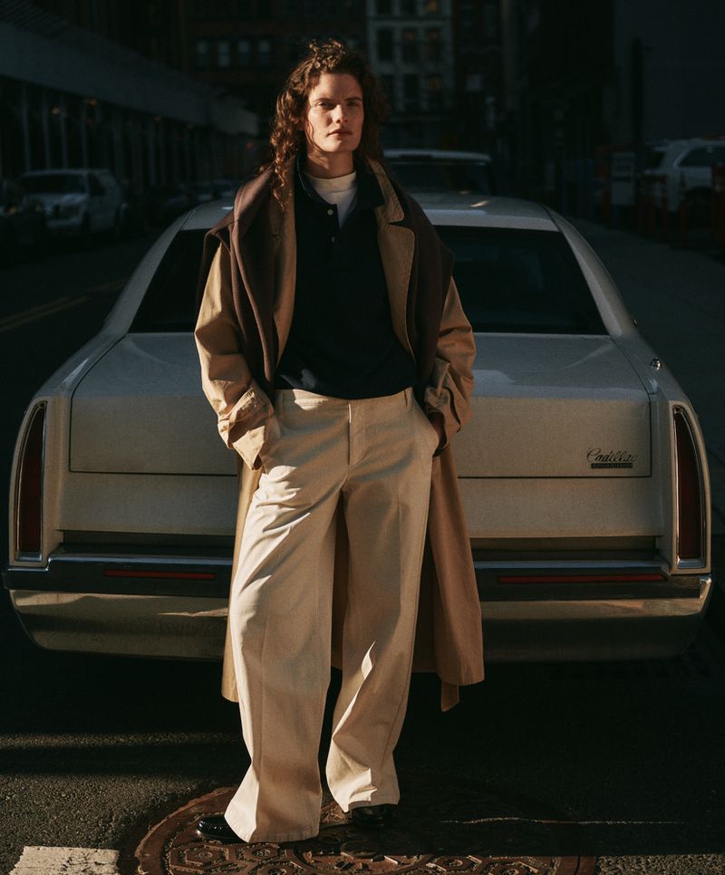 Adult with long curly hair, posing next to a vintage Cadillac, wearing cream-colored pants and a beige coat on a city street.