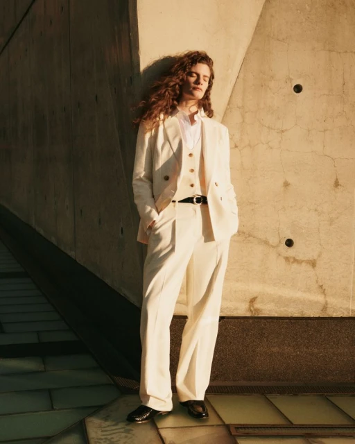 Woman in an ivory suit standing against a textured architectural background with sunlight accentuating her curly brown hair.
