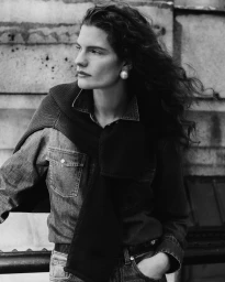 Model with long dark wavy hair wearing a denim shirt and a sweater tied over her shoulders outdoors.