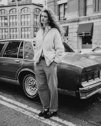 Model standing outdoors next to a parked car wearing a cardigan and trousers in a monochrome editorial photograph
