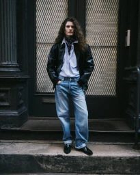 Model with long curly hair wearing a black leather jacket, light blue shirt, and jeans standing in front of a doorway.