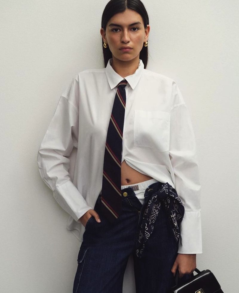 Portrait of a woman in a white oversized shirt, tie, and dark blue jeans, holding a black bag against a neutral wall.