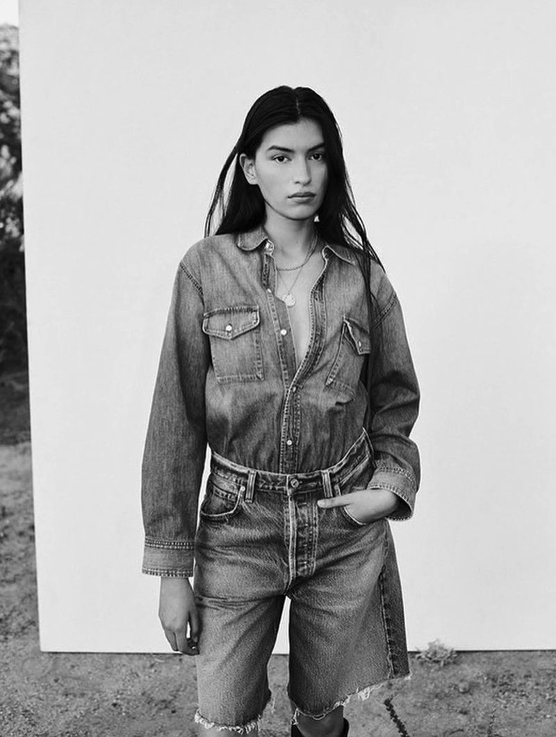 Woman poses in black and white, wearing a denim shirt and jean shorts against a bright background in an outdoor environment.