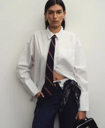 Model in white button-down shirt, striped tie, and dark denim jeans standing against a plain white wall