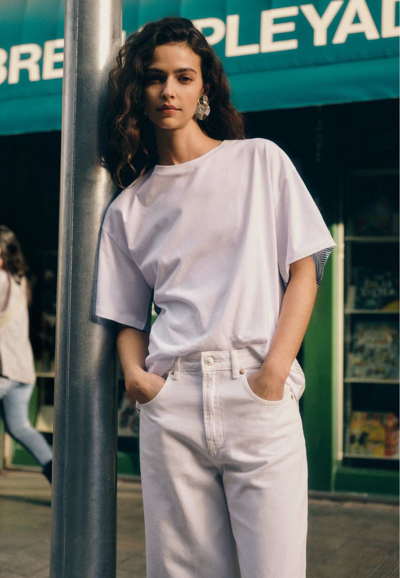 A woman in casual white clothing poses in an urban setting, leaning against a pole near a storefront for a fashion-forward, lifestyle image.
