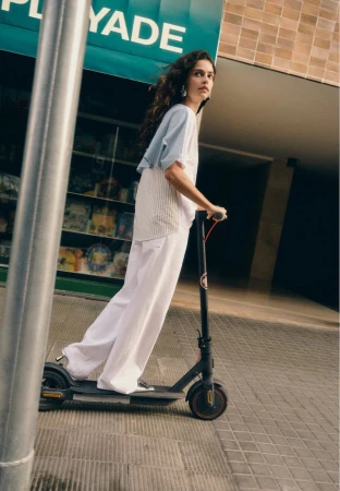 A young woman riding a scooter on a city sidewalk in casual summer clothing, captured from a low angle, with natural light.
