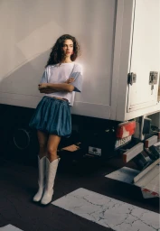 Model with long dark curly hair wearing a white t-shirt, blue bubble skirt, and white boots leaning against a truck.