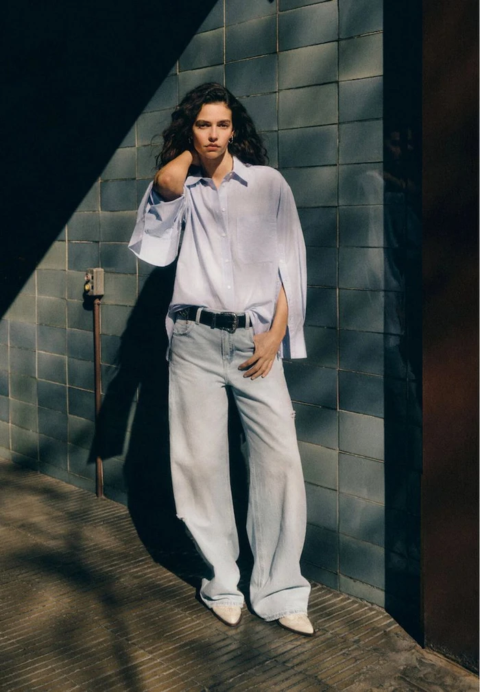 Model standing outdoors wearing a light blue button-down shirt and wide-leg jeans against a dark tiled wall under sunlight.