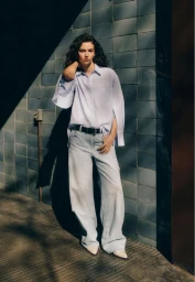 Model standing outdoors wearing a light blue button-down shirt and wide-leg jeans against a dark tiled wall under sunlight.