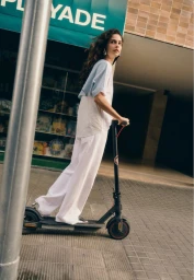 Woman with long dark hair riding an electric scooter on a sidewalk past a shopfront for Stradivarius