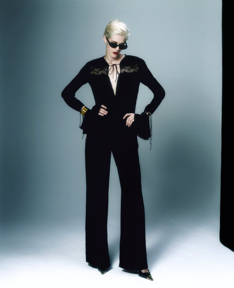 An adult female fashion model poses indoors, wearing a black lace top and wide leg pants, accessorized with cuffs and sunglasses.