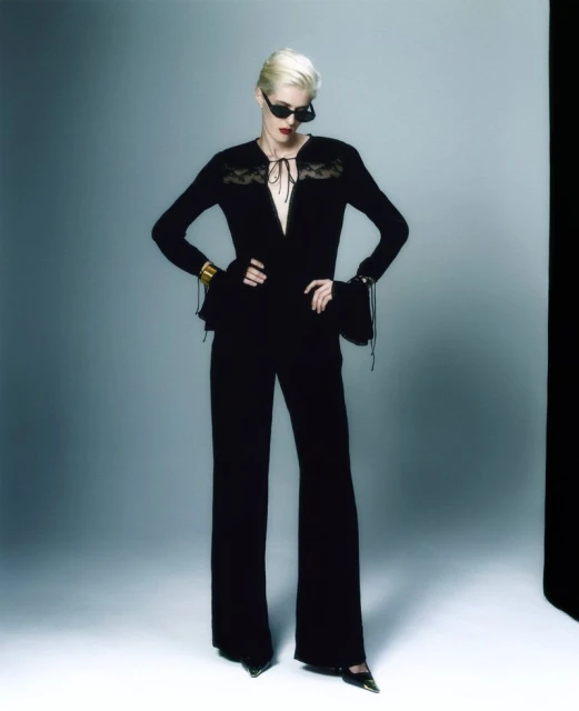 An adult female fashion model poses indoors, wearing a black lace top and wide leg pants, accessorized with cuffs and sunglasses.