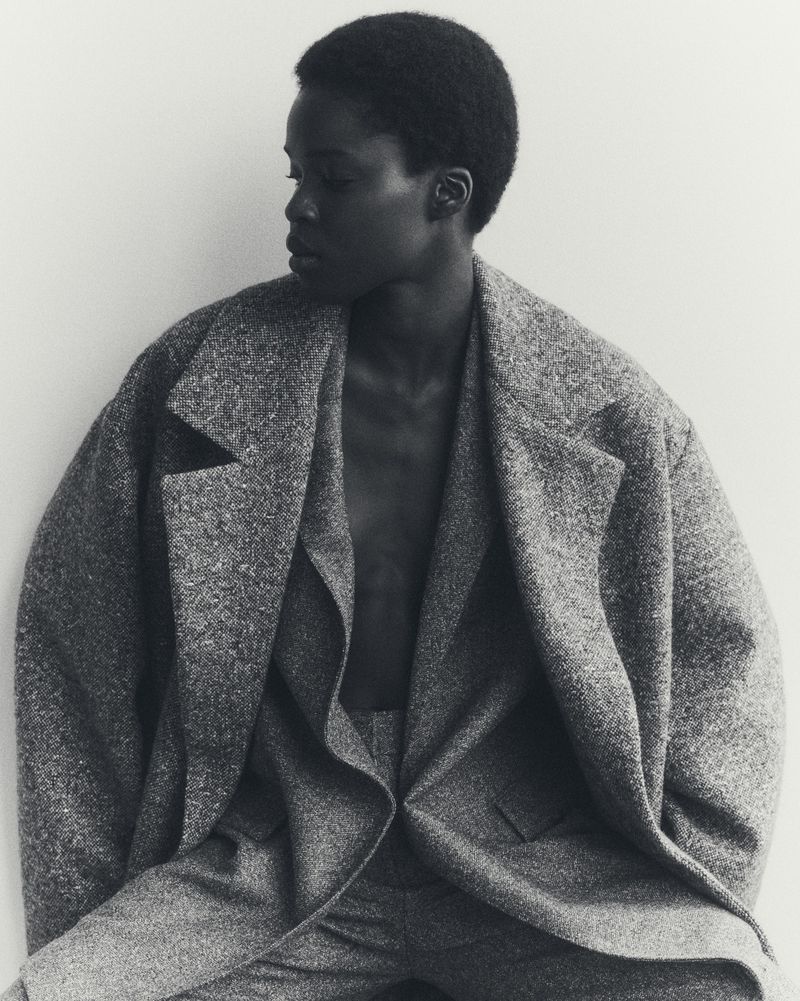 Monochrome studio portrait of an African woman in a grey textured oversized blazer, looking off-camera, neutral expression, white backdrop.