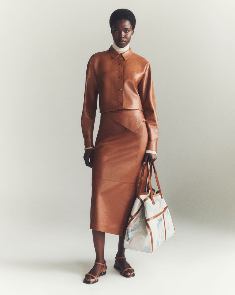 Full-length fashion shot of an African-American woman in a brown leather skirt suit with a patterned tote bag, posing against a neutral backdrop.