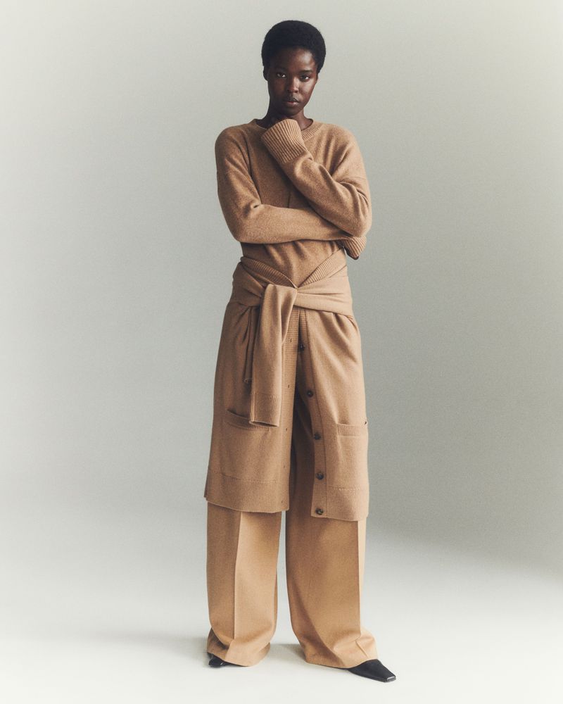 Full-length studio portrait of a slender Black female model in a tan knit sweater and pants with a tied sweater around the waist.