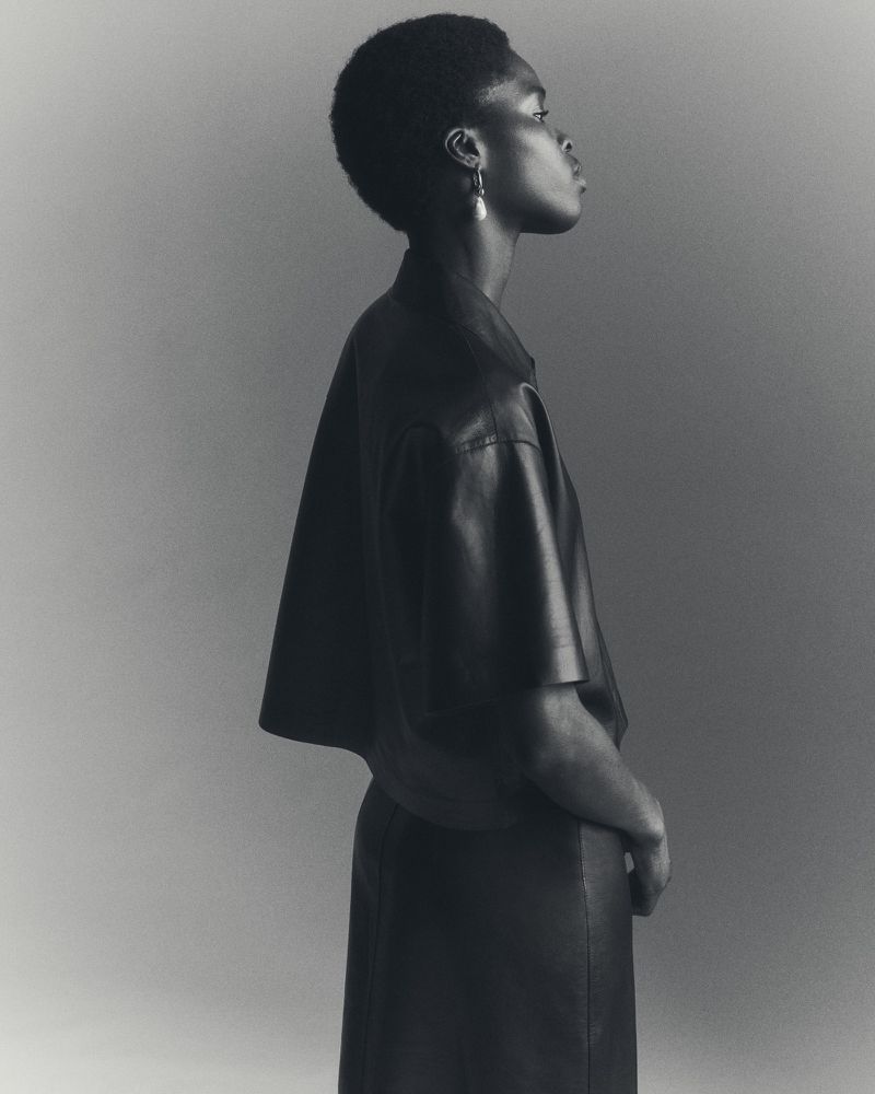 Black and white profile shot of a woman with short hair wearing a dark top and skirt. The studio lighting highlights her features.