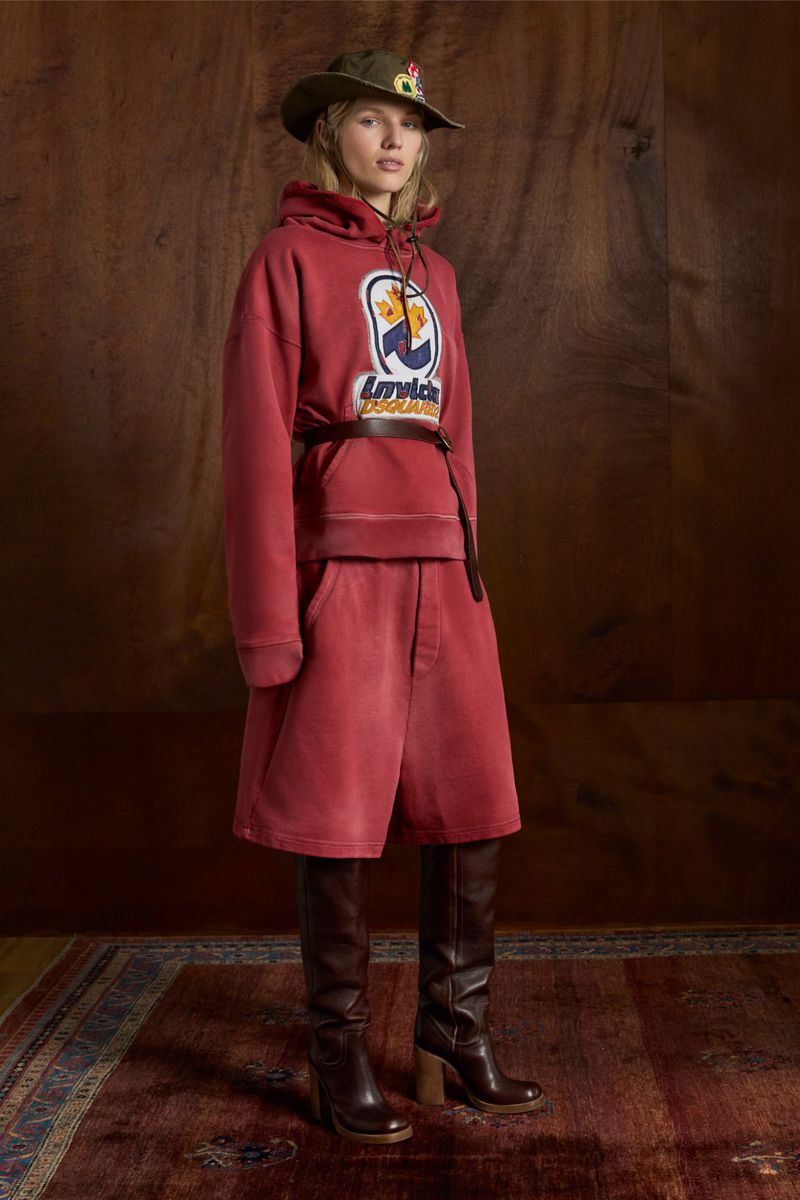 Woman in a red hoodie and shorts, paired with heeled boots and a bucket hat, poses in front of a wood-paneled wall and vintage rug.