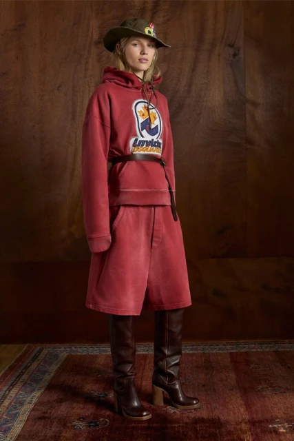 Woman in a red hoodie and shorts, paired with heeled boots and a bucket hat, poses in front of a wood-paneled wall and vintage rug.