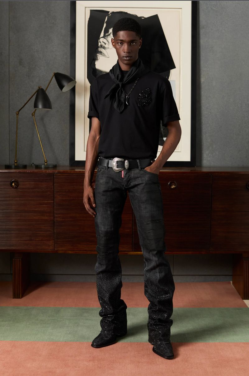 A stylish young man poses indoors wearing a black t-shirt, jeans, boots, and accessories. A wooden sideboard is in the background.