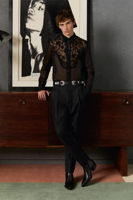 A young adult man poses indoors near a wooden console table with a painting, wearing a black sheer and lace shirt with black pants and boots.