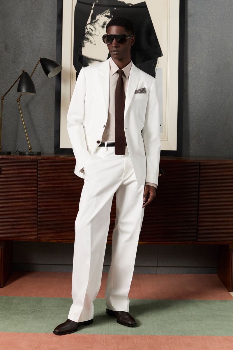 Man in white suit and sunglasses stands indoors next to cabinet and framed art, posing for a full-length fashion portrait.