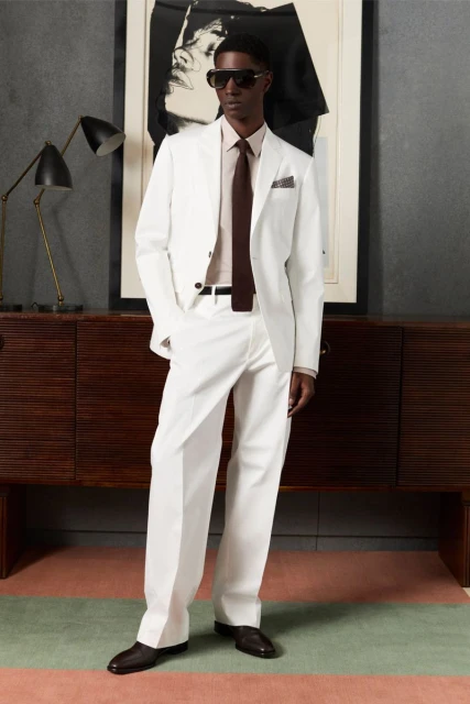 Man in white suit and sunglasses stands indoors next to cabinet and framed art, posing for a full-length fashion portrait.