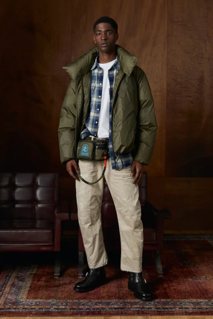 A young adult male model stands in a photo studio. He wears an olive puffer jacket, plaid shirt, beige pants, black boots, and a waist bag.