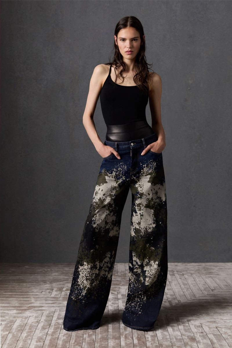 A woman stands in a studio wearing a black tank top and wide-leg jeans with a dark and light mottled wash.