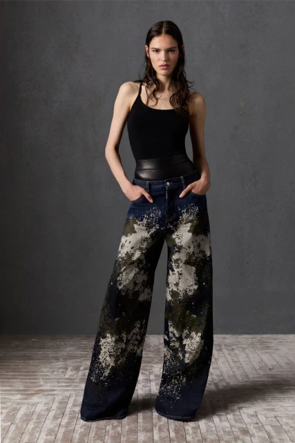 A woman stands in a studio wearing a black tank top and wide-leg jeans with a dark and light mottled wash.