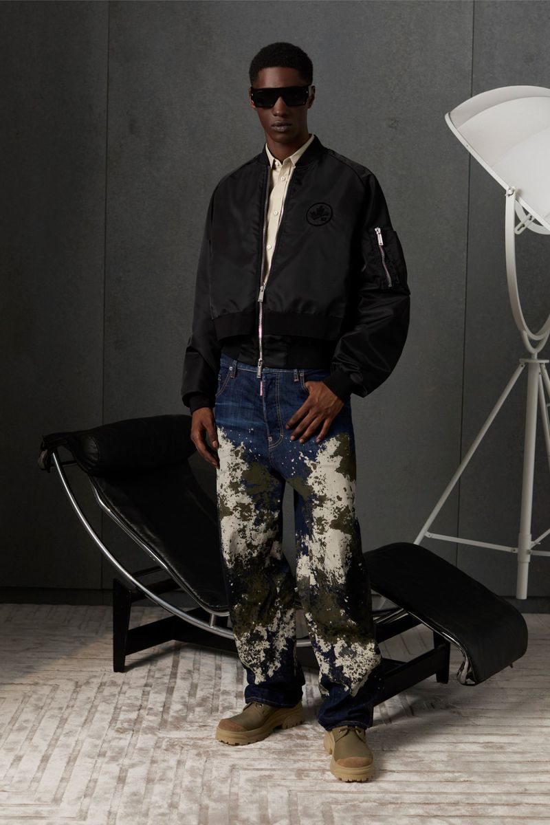 A man in a black bomber jacket, digitally patterned jeans, and sunglasses poses in a neutral setting with studio lighting.