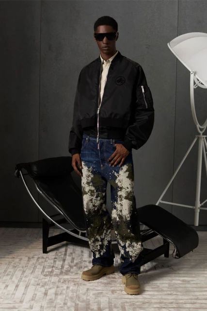 A man in a black bomber jacket, digitally patterned jeans, and sunglasses poses in a neutral setting with studio lighting.