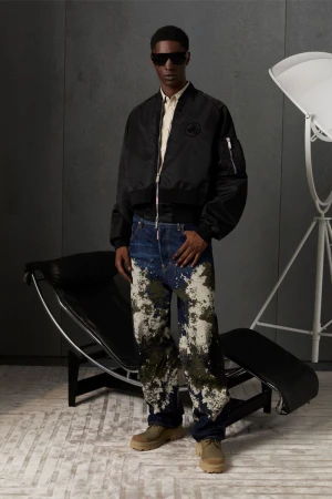 A man in a black bomber jacket, digitally patterned jeans, and sunglasses poses in a neutral setting with studio lighting.