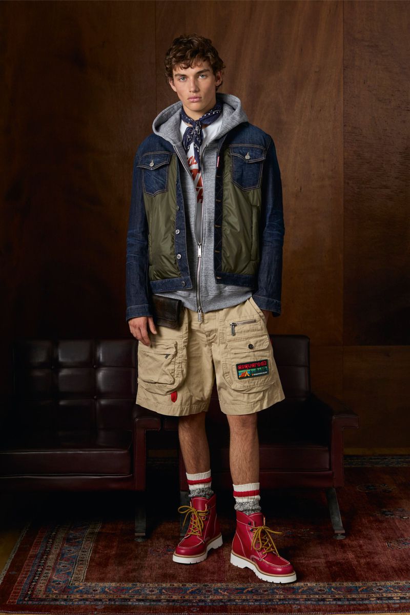 Full-length shot of a young adult man in a layered outfit including a jacket, hoodie, shorts, boots, and socks in an indoor studio.