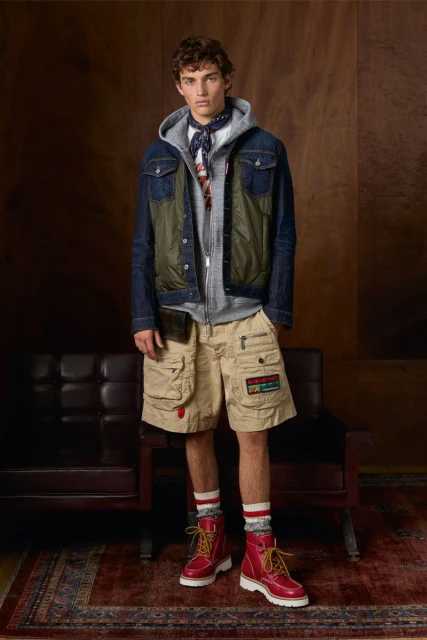 Full-length shot of a young adult man in a layered outfit including a jacket, hoodie, shorts, boots, and socks in an indoor studio.