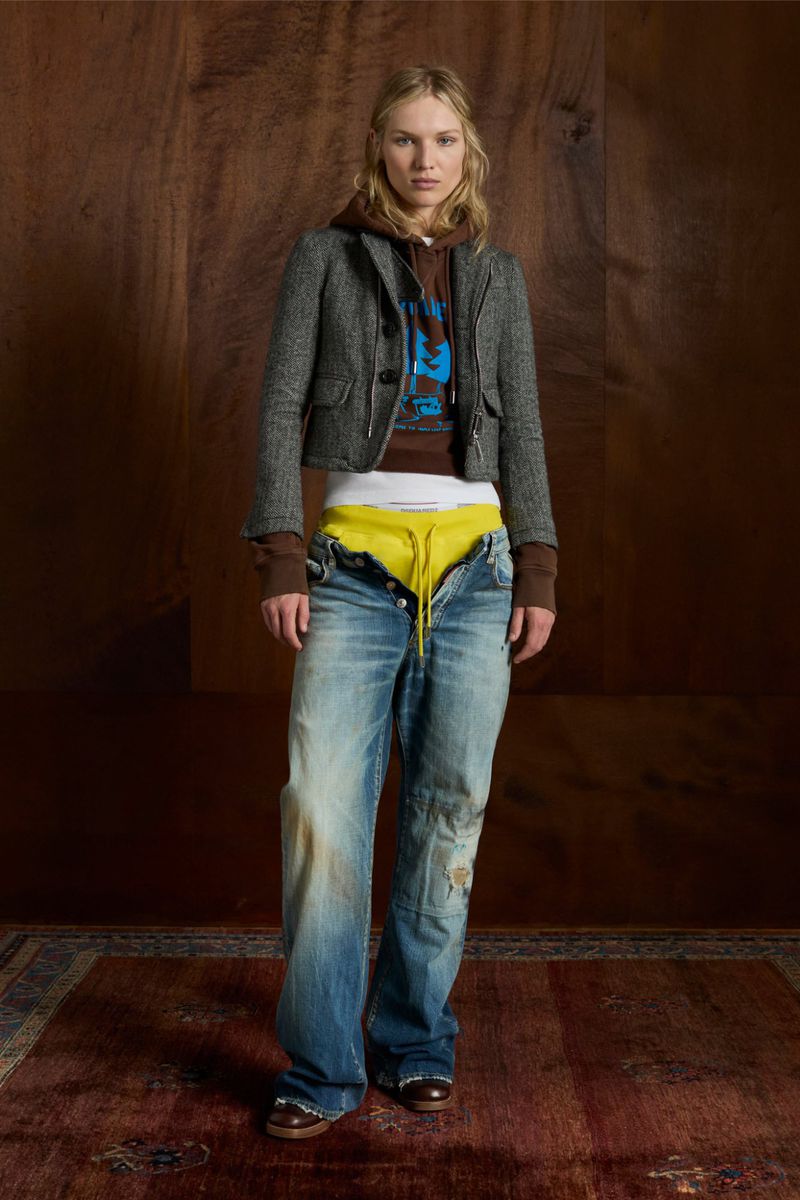 Woman in fashionable pose wearing a gray tweed jacket, brown hoodie, and ripped jeans stands against a wood backdrop and patterned rug.